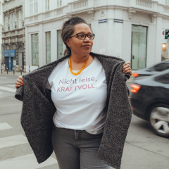 Person steht auf einer Straßenecke und hält einen offenen Mantel, darunter ein weißes T-Shirt mit dem Schriftzug 'Nicht leise, KRAFTVOLL.'